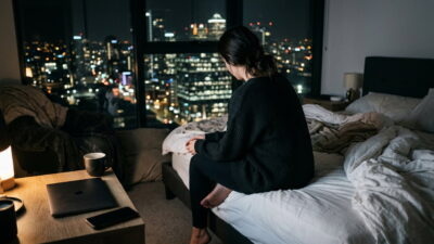 Woman sits on a messy bed at night, looking out a window at a city skyline with lit buildings and streetlights outside the glow of the urban night.